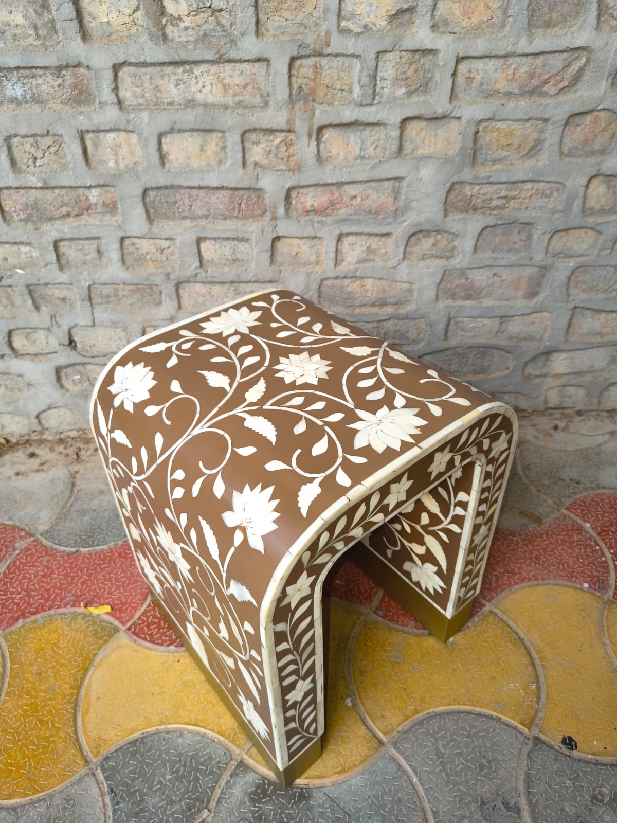 A small upholstered stool with a curved back and seat, covered in brown fabric with a white floral vine pattern, photographed outdoors against a stone wall and patterned tile floor.