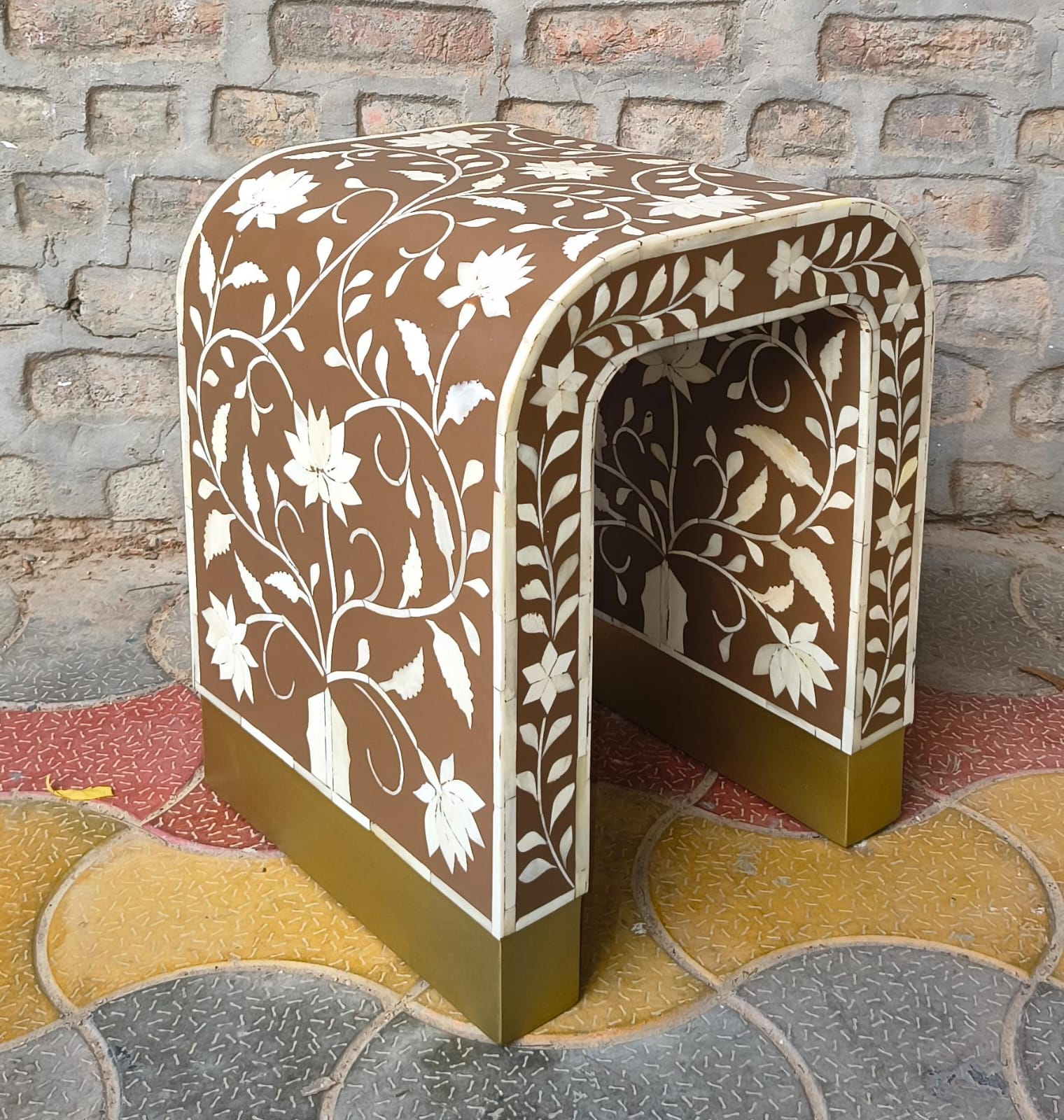 A small arched bench or stool with a brown base decorated with white floral inlay, set against a brick wall and patterned tile floor.