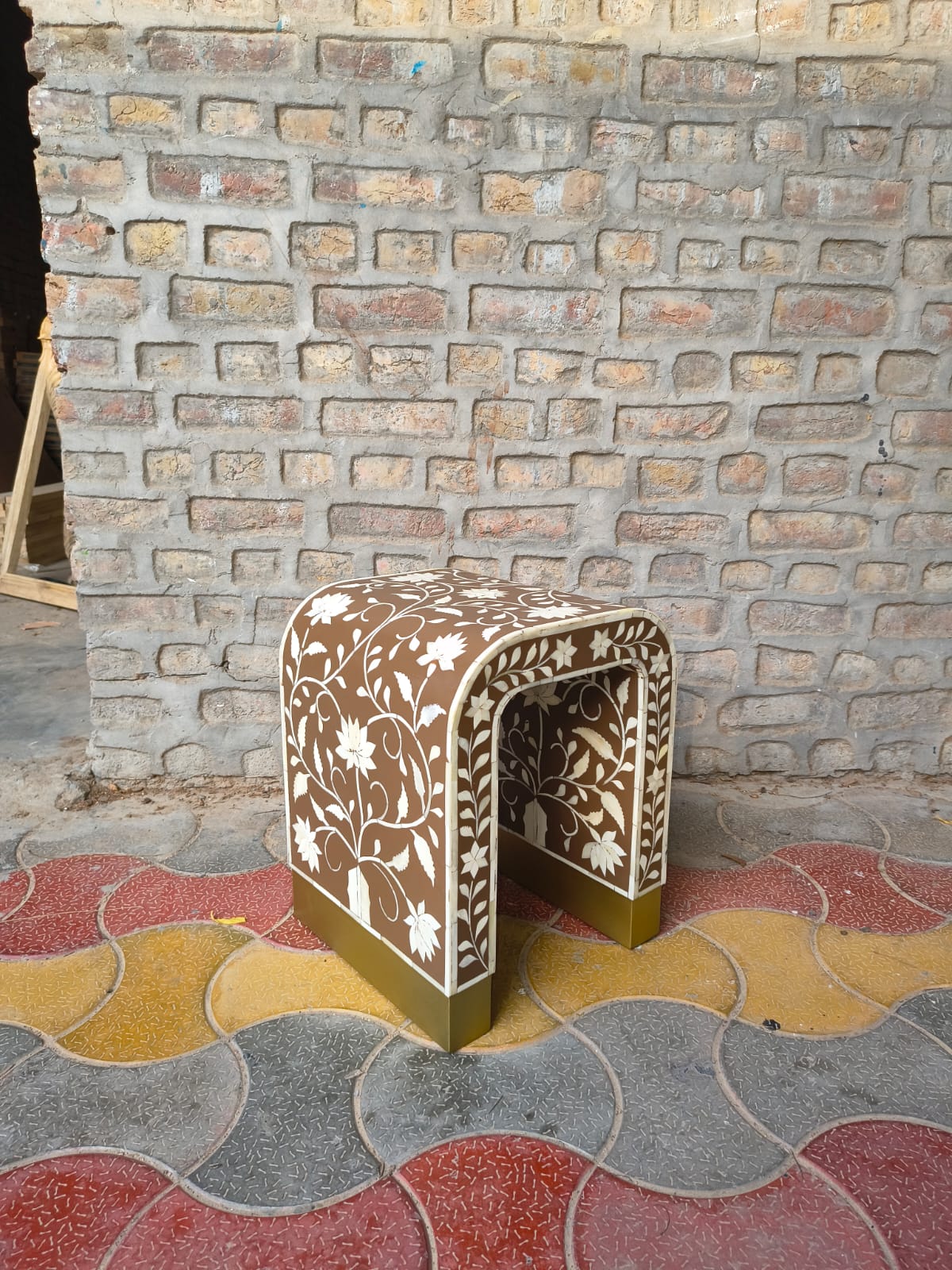 A small decorative stool with a brown and beige floral inlay pattern, set on a patterned red, yellow, and gray stone floor against a rustic brick wall.