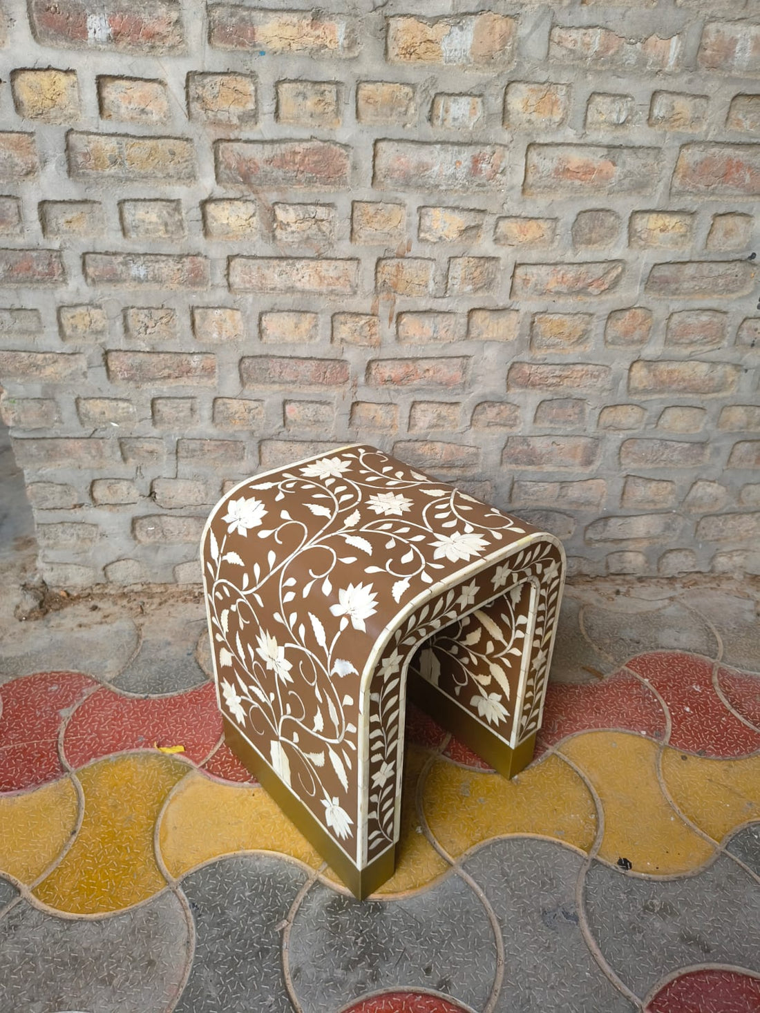 A small arched bench or stool with a brown base decorated with white floral inlay, set against a brick wall and patterned tile floor.